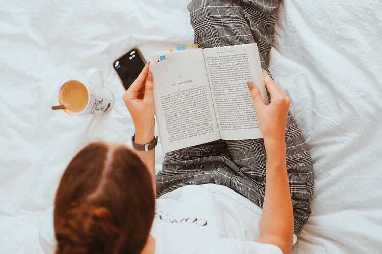 Mulher lendo um livro na cama com uma xícara de café ao lado.