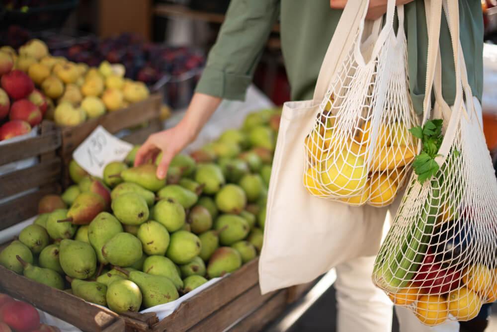Mulher comprando frutas com sacolas reutilizáveis.
